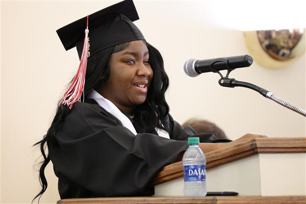 A graduate from the Alabama School for the Blind speaks into a microphone from behind a podium at Baccalaureate. She is wearing a black cap and gown with a red and white tassel. 